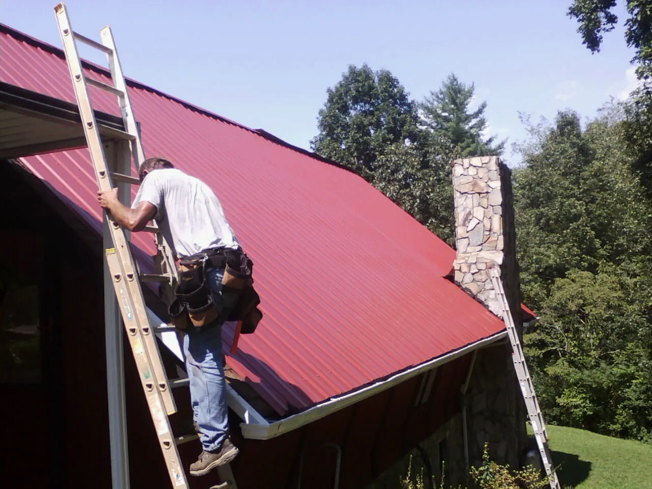 Roofer on a ladder beside a red metal roof and stone chimney — Soffit & Fascia Repair by Roofing Brothers in Center Cross